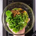 A hand holds a glass bowl containing chopped cilantro, parsley, red pepper flakes, oil, and seasonings above a black grill pan.