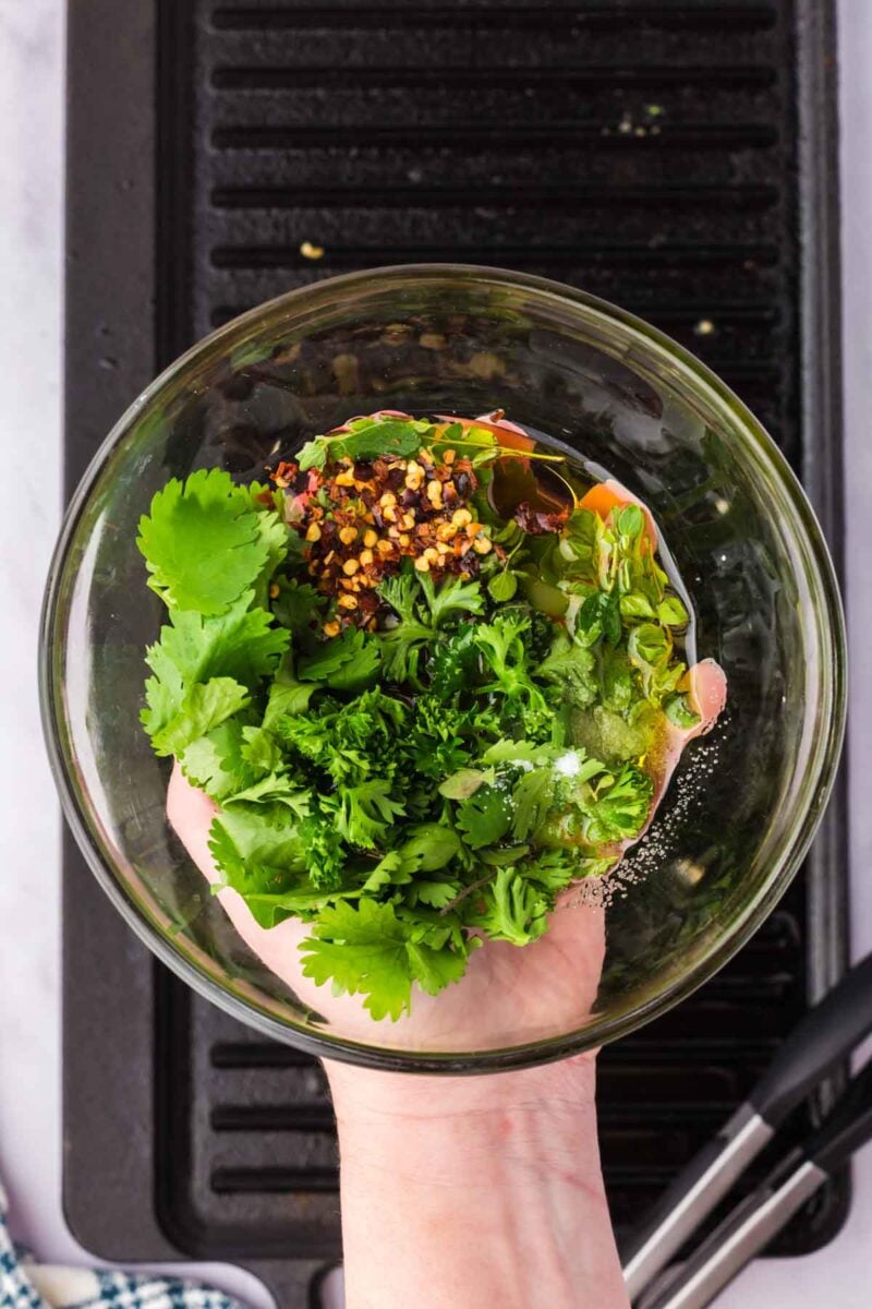 A hand holds a glass bowl containing chopped cilantro, parsley, red pepper flakes, oil, and seasonings above a black grill pan.