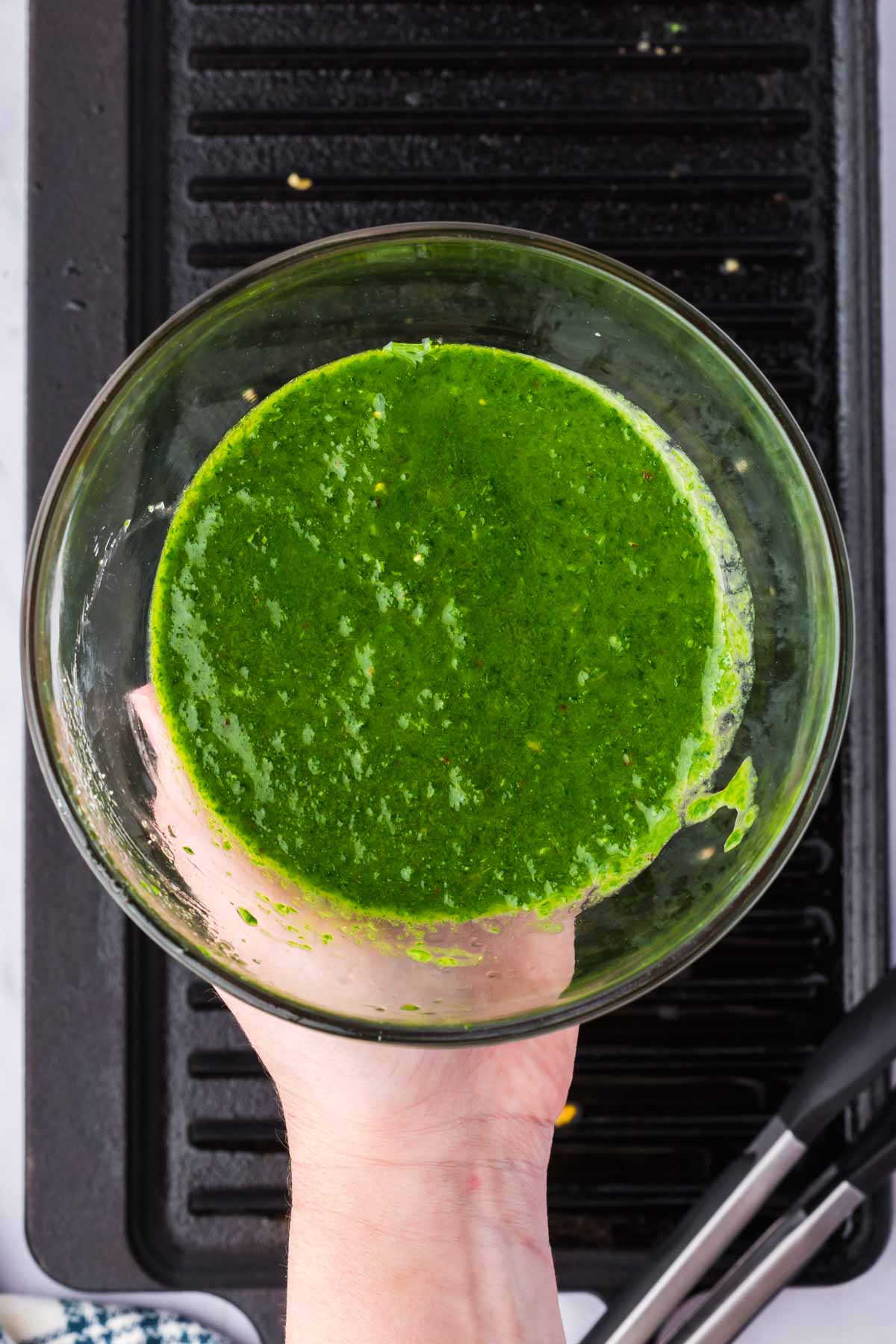 A hand holds a glass bowl filled with bright green sauce above a black grill pan.