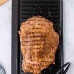 Grilled flank steak with char marks sits on a black grill pan, next to a pair of metal tongs, a cutting board with herbs, and a bowl of green sauce.