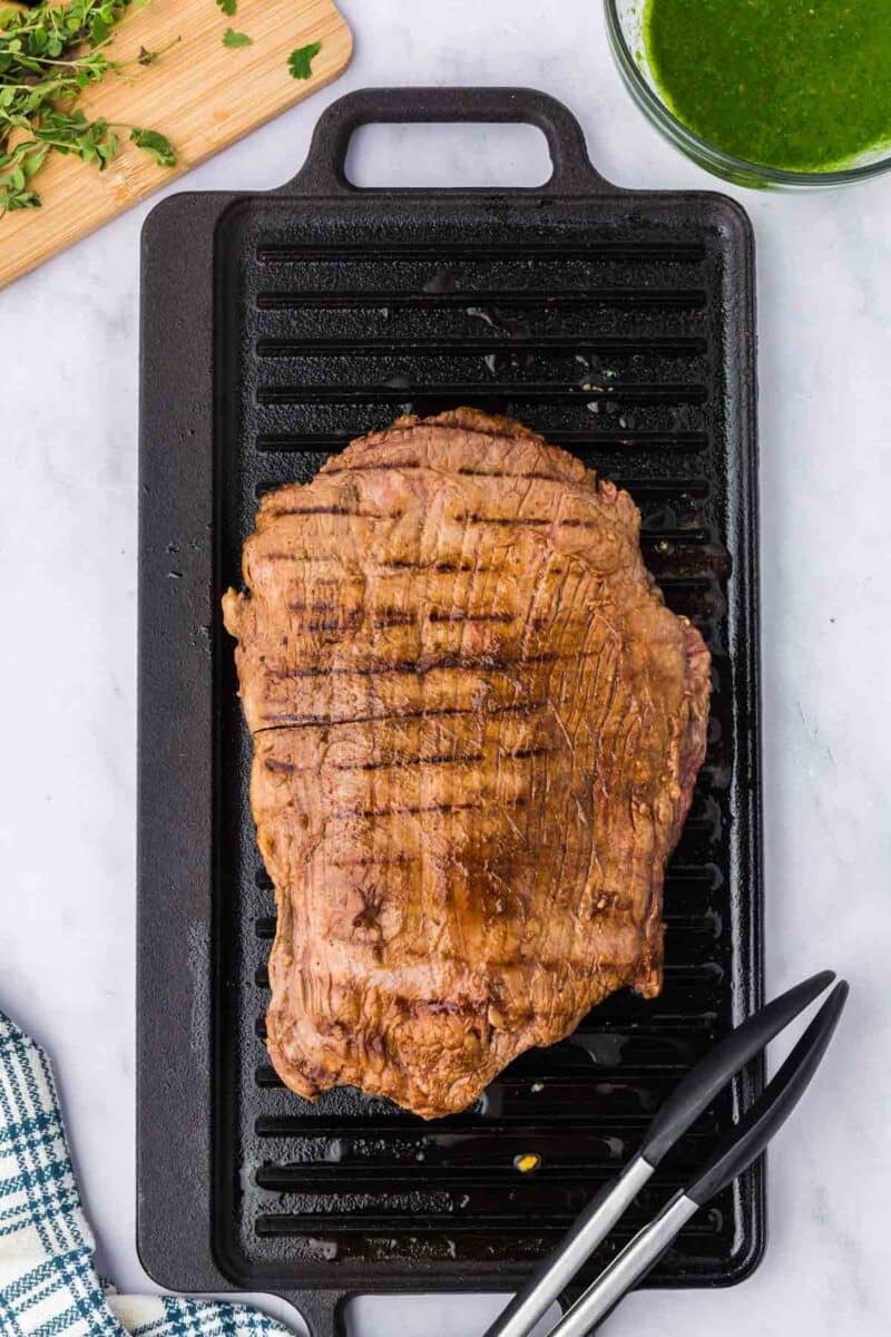 Grilled flank steak with char marks sits on a black grill pan, next to a pair of metal tongs, a cutting board with herbs, and a bowl of green sauce.