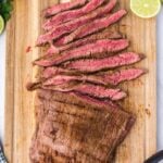 Sliced grilled flank steak served on a wooden cutting board with lime halves, fresh herbs, and metal tongs nearby.