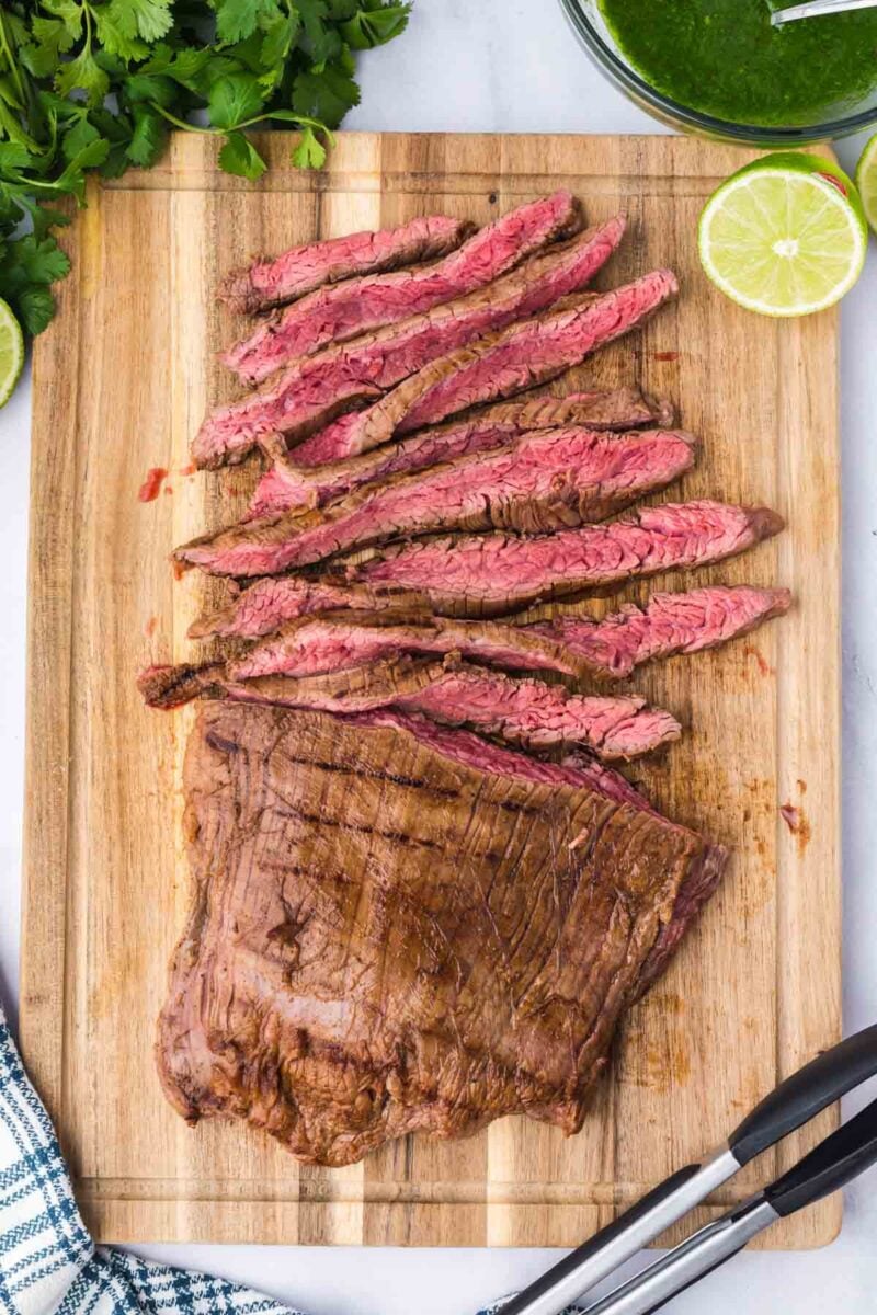 Sliced grilled flank steak served on a wooden cutting board with lime halves, fresh herbs, and metal tongs nearby.