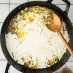 A large black pot filled with a creamy liquid and seasonings, with a wooden spoon resting on the side; salt, pepper, and a lemon juicer are nearby on a tiled surface.