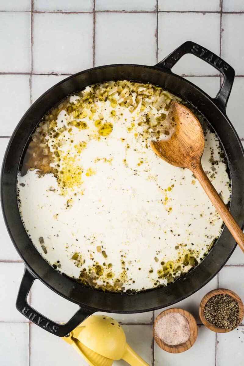A large black pot filled with a creamy liquid and seasonings, with a wooden spoon resting on the side; salt, pepper, and a lemon juicer are nearby on a tiled surface.
