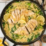 A skillet containing creamy orzo with spinach and asparagus, topped with sliced cooked chicken breast and garnished with lemon slices.