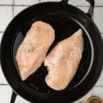 Two raw chicken breasts seasoned with salt and pepper in a black cast iron pan, with small wooden bowls of salt and pepper beside the pan on a tiled surface.