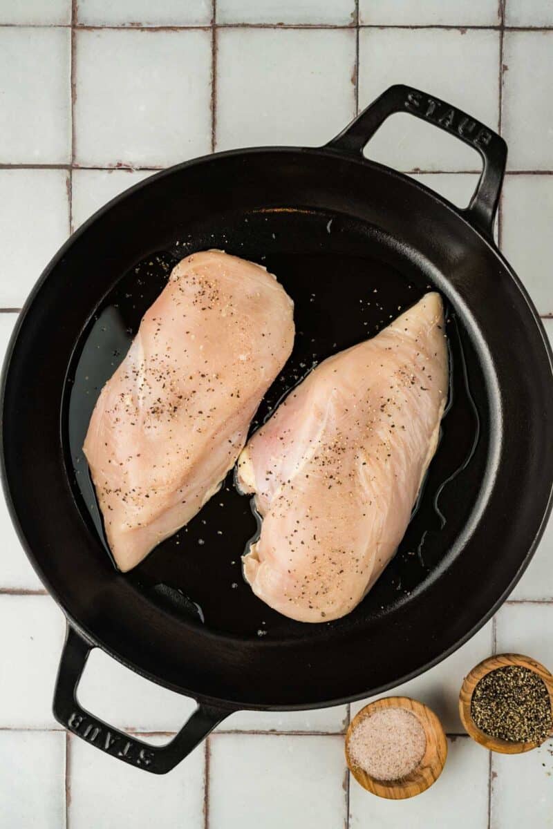 Two raw chicken breasts seasoned with salt and pepper in a black cast iron pan, with small wooden bowls of salt and pepper beside the pan on a tiled surface.