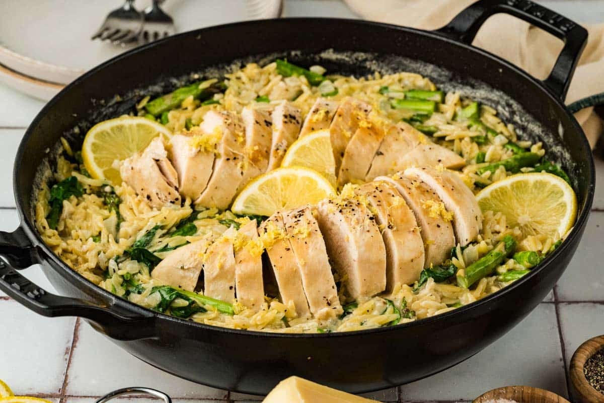 Sliced cooked chicken breast on a bed of creamy orzo with spinach and asparagus, garnished with lemon slices in a black skillet.