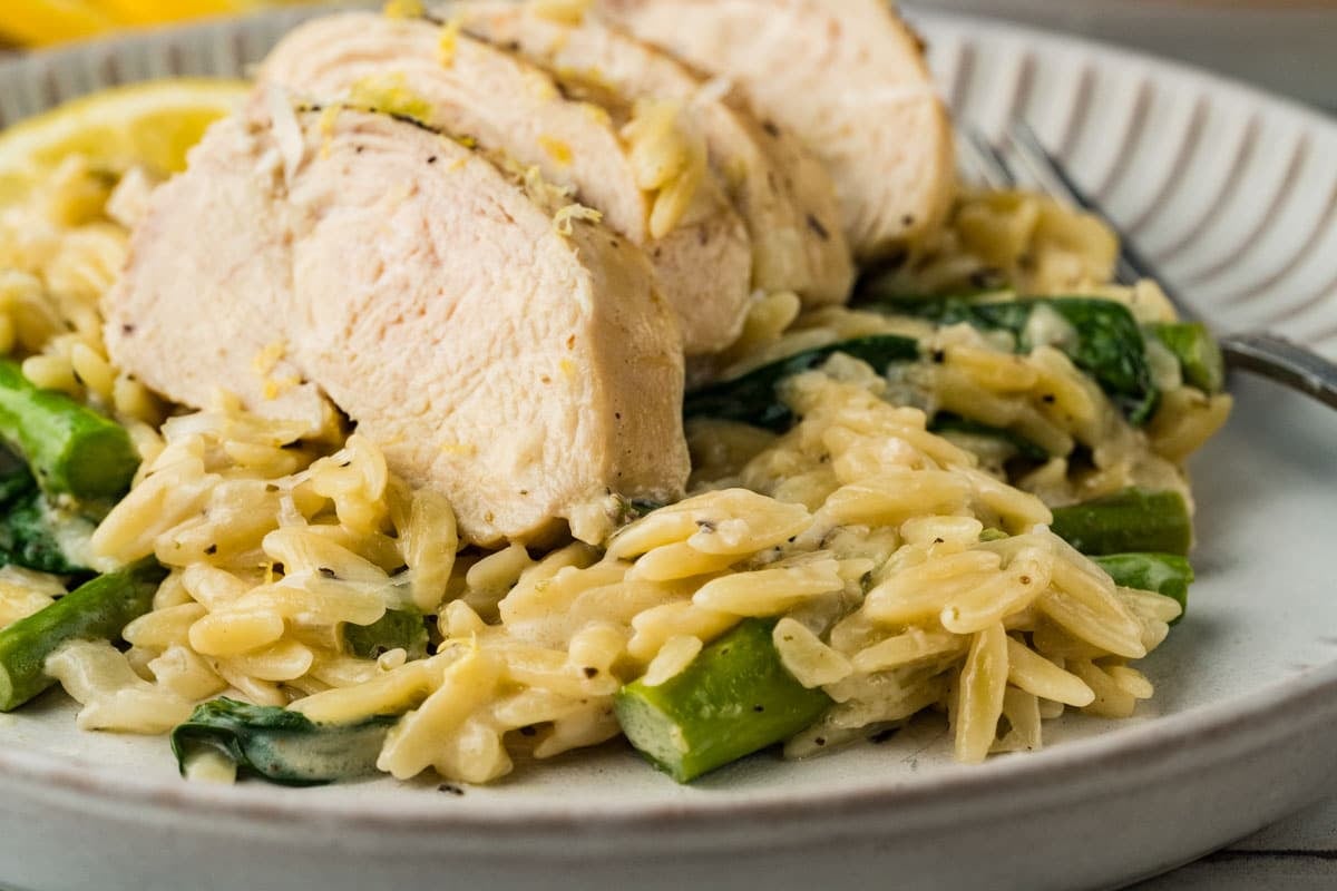 Sliced cooked chicken breast served over creamy orzo pasta with spinach and asparagus on a white plate.