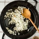 Chopped onions cooking in a black cast iron pan with oil, stirred by a wooden spoon; small bowls of salt and pepper are nearby.