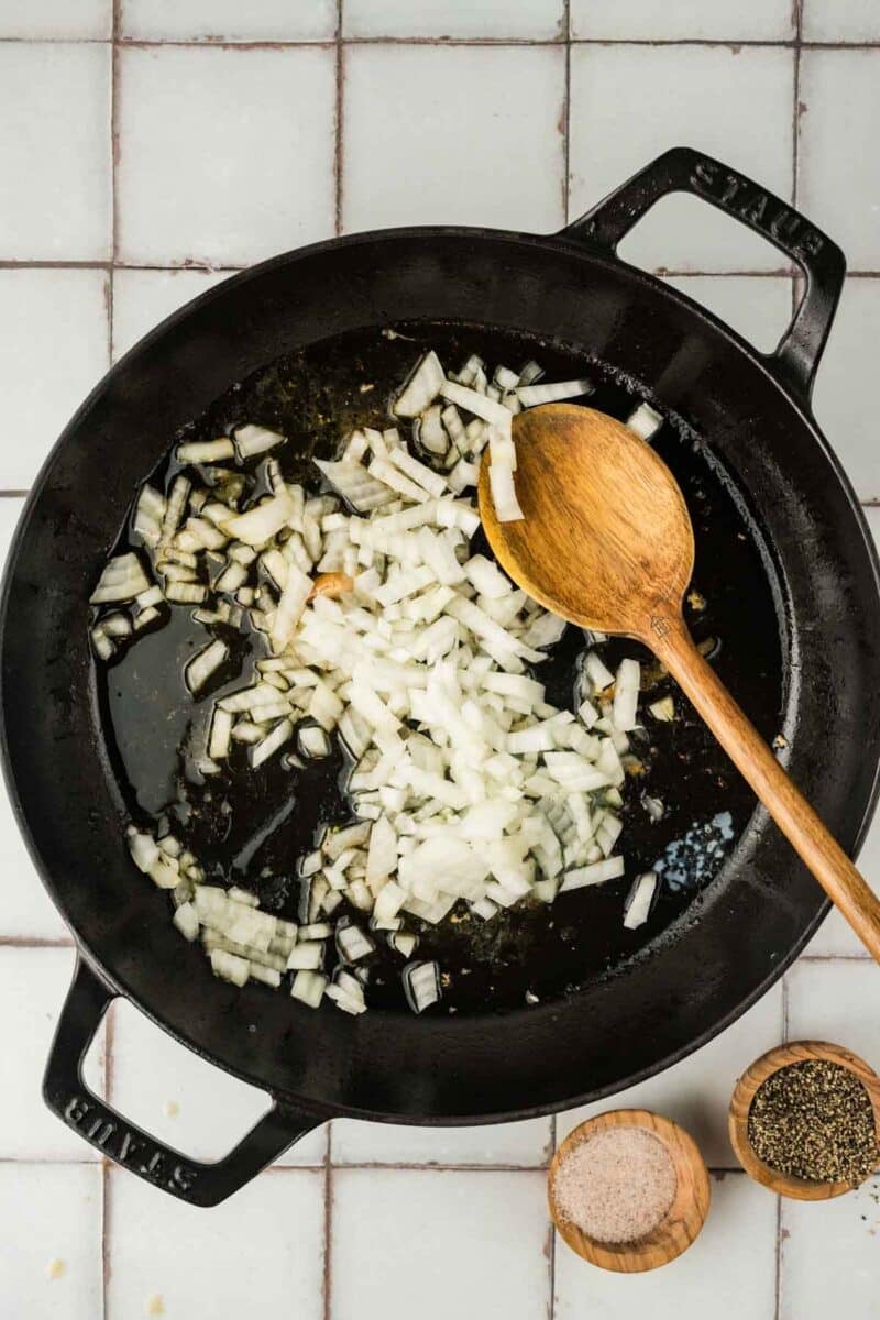 Chopped onions cooking in a black cast iron pan with oil, stirred by a wooden spoon; small bowls of salt and pepper are nearby.