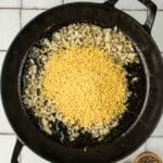 A black skillet with sautéed onions and dry orzo pasta, next to small wooden bowls of salt and black pepper on a tiled surface.