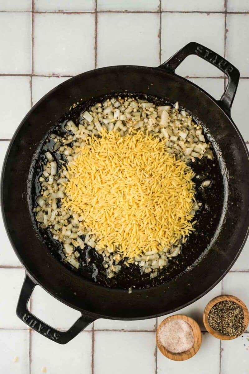 A black skillet with sautéed onions and dry orzo pasta, next to small wooden bowls of salt and black pepper on a tiled surface.