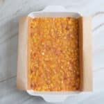 A white baking dish lined with parchment paper is filled with an uncooked peach mixture on a light marble surface.