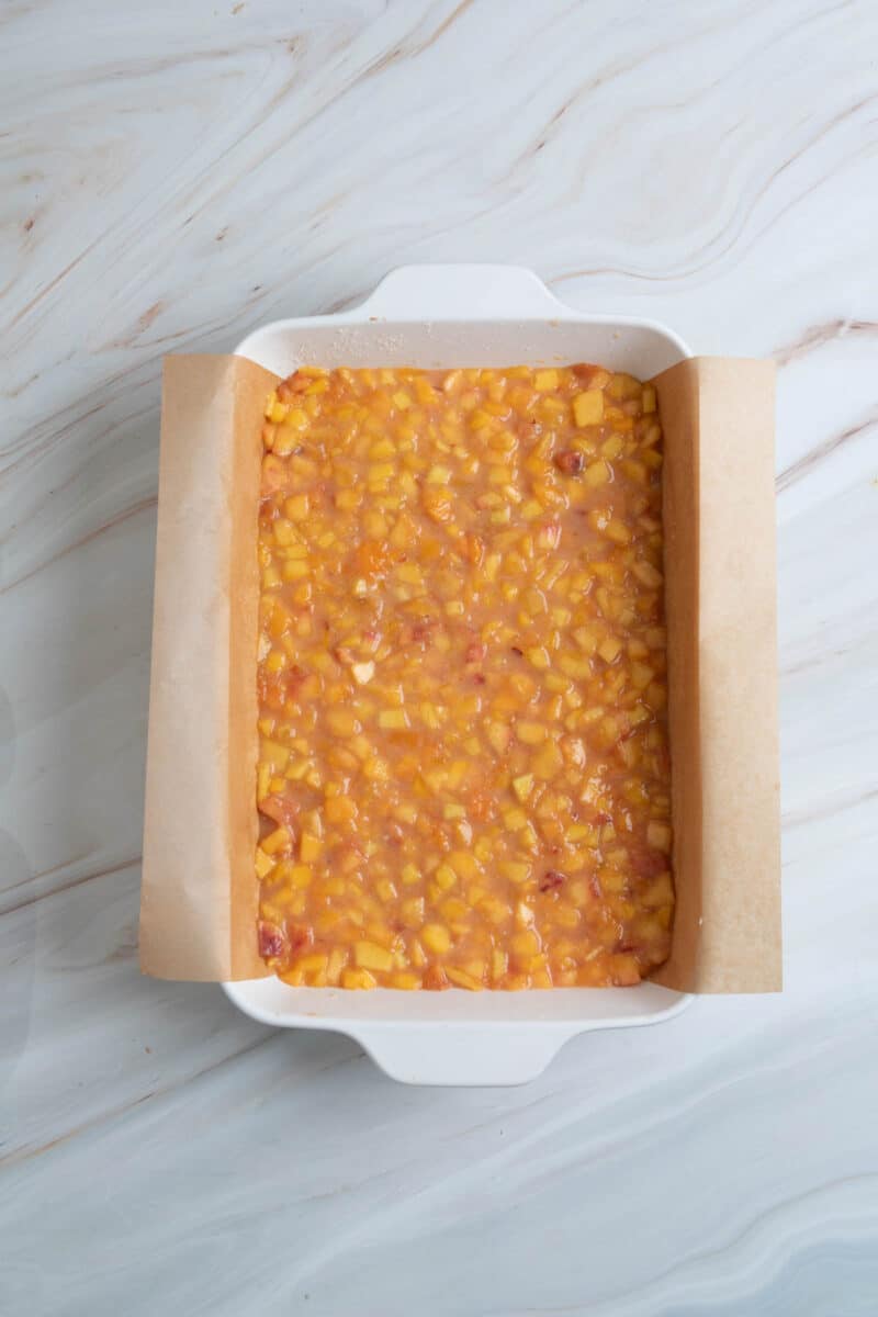 A white baking dish lined with parchment paper is filled with an uncooked peach mixture on a light marble surface.