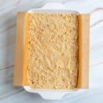 A baking dish lined with parchment paper is filled with a layer of crumbly, light brown mixture on a marble surface.