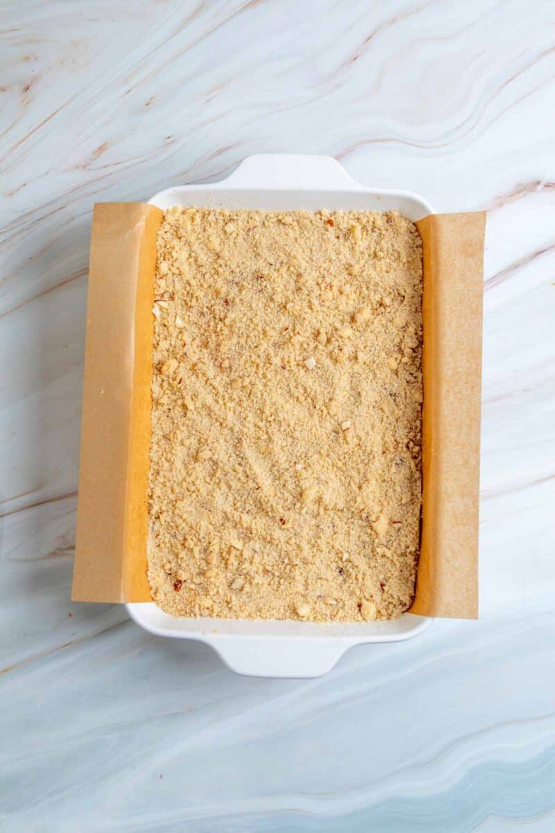 A baking dish lined with parchment paper is filled with a layer of crumbly, light brown mixture on a marble surface.