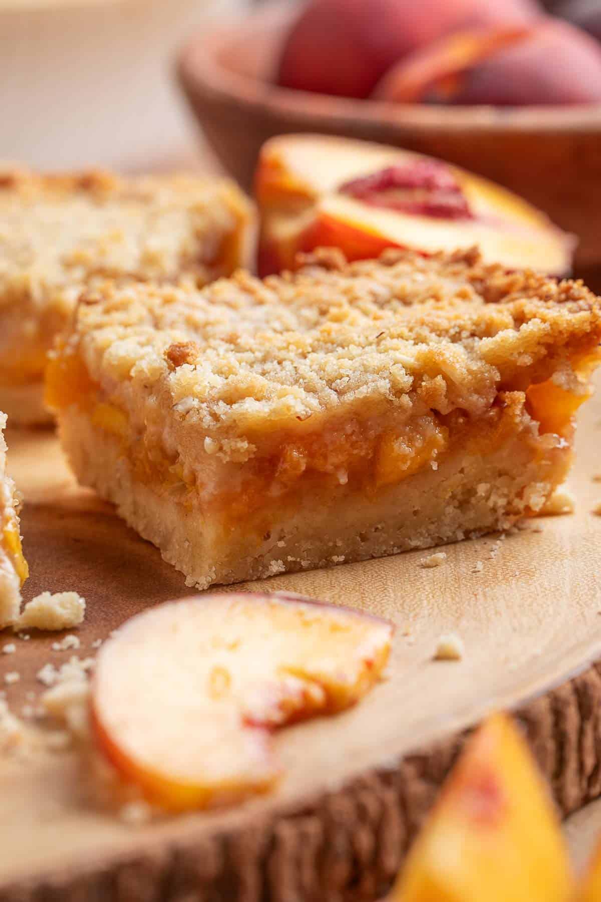 A close-up of a peach crumb bar on a wooden surface, with fresh peach slices and whole peaches in the background.