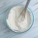 A glass bowl filled with flour and a metal whisk resting inside, placed on a white marble surface.