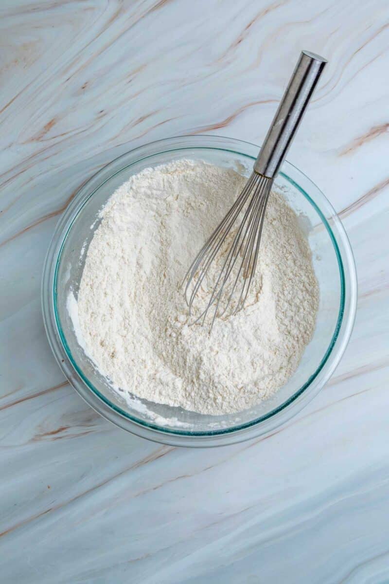 A glass bowl filled with flour and a metal whisk resting inside, placed on a white marble surface.