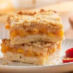 Two crumb-topped peach bars are stacked on a white plate, with a peach slice beside them.