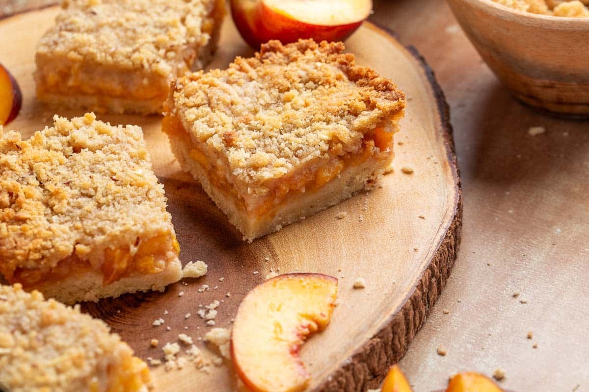 Four peach crumb bars on a wooden platter with sliced peaches and crumbs scattered around.