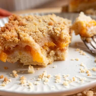 A slice of peach barsย on a white plate with a fork and peach slices, showing a crumbly topping and peach filling.