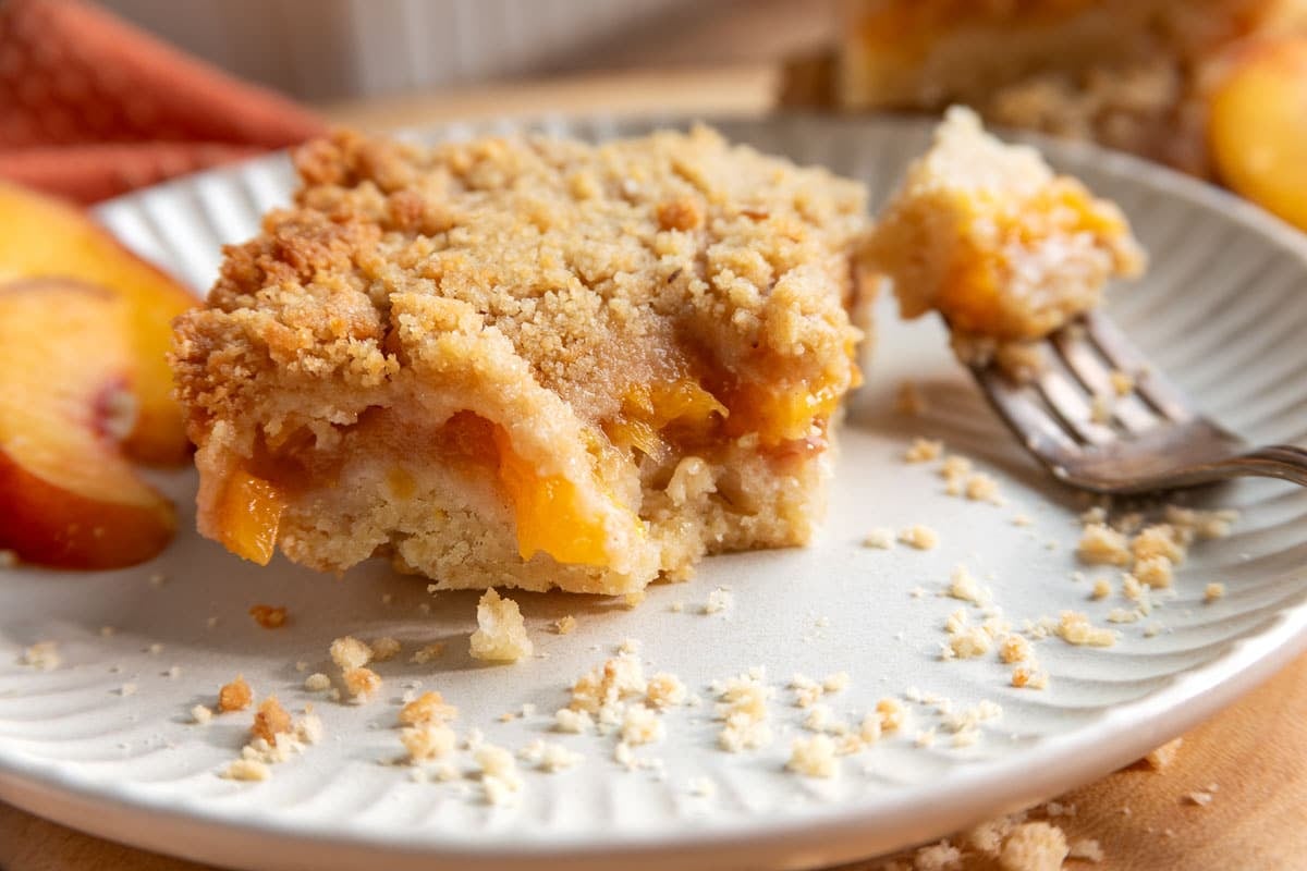 A slice of peach bars on a white plate with a fork and peach slices, showing a crumbly topping and peach filling.