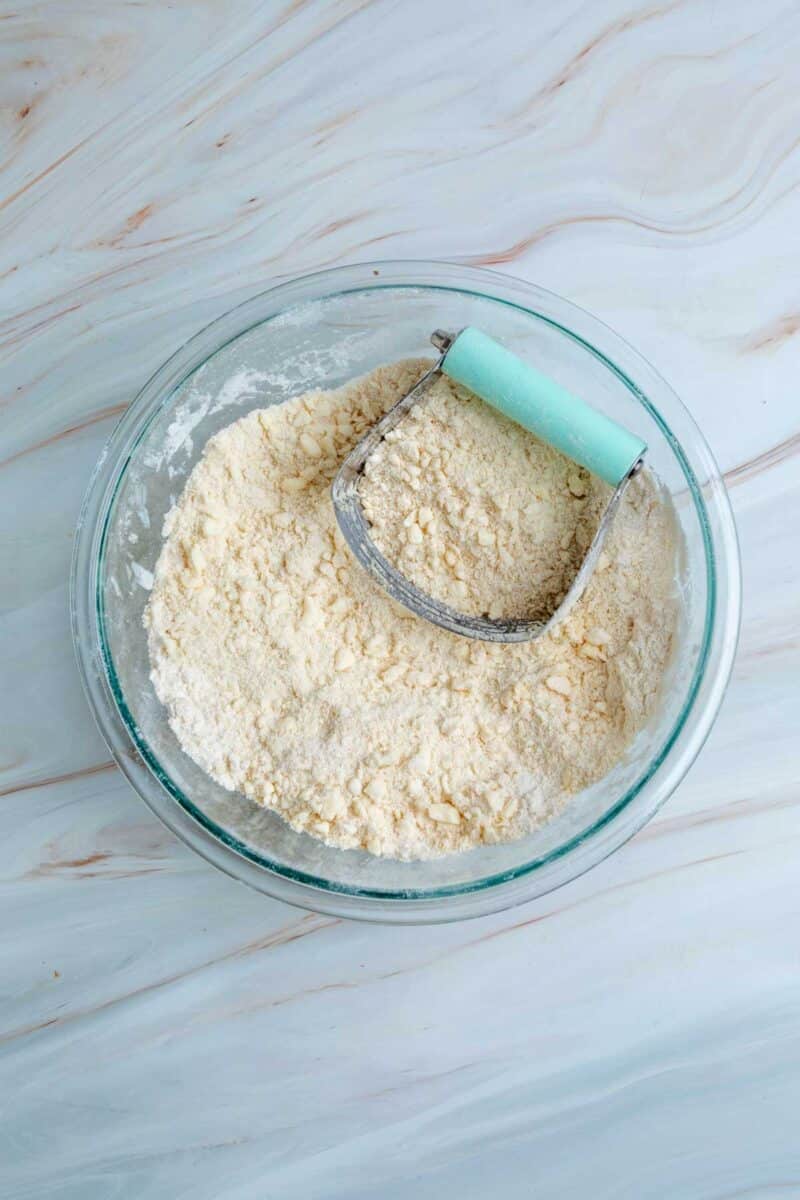 A glass bowl with flour and butter being mixed using a pastry cutter, on a light marble surface.