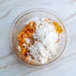 A clear glass bowl with chopped peaches and a large amount of flour and sugar on top, placed on a white marble surface.