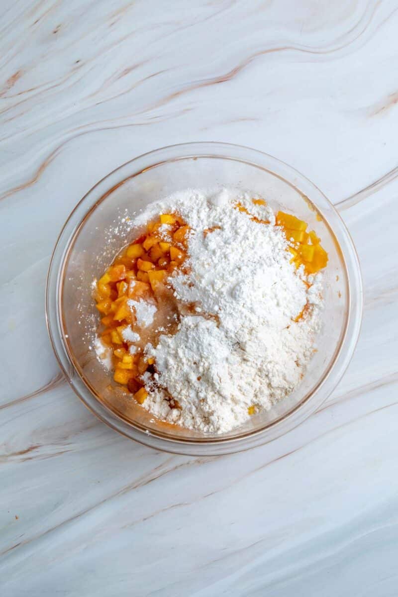 A clear glass bowl with chopped peaches and a large amount of flour and sugar on top, placed on a white marble surface.