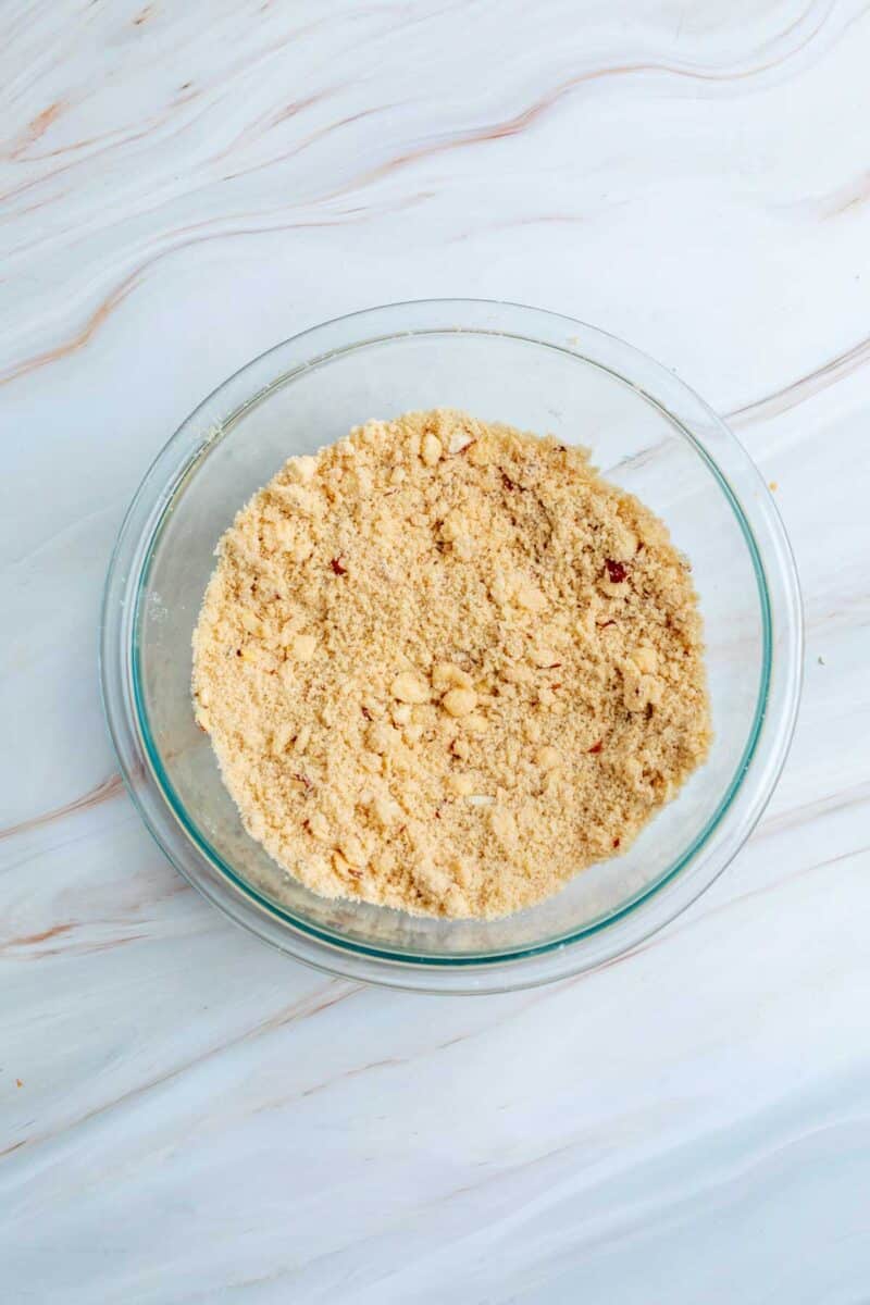 A clear glass bowl filled with crumbly, light brown mixture sits on a white marbled surface.