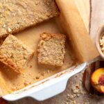 A white baking dish with parchment paper holds two crumbly peach bars. One piece is separated, and sliced peaches and crumbs are nearby on the table.
