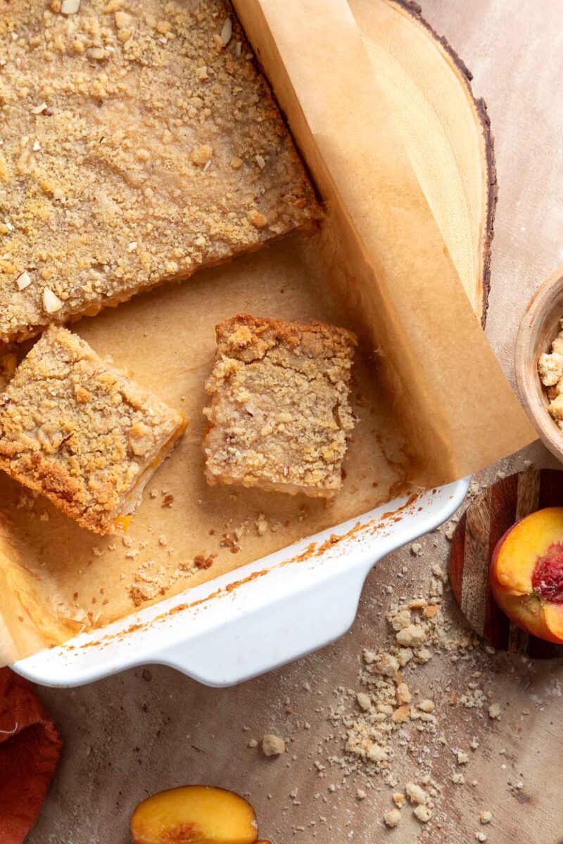 A white baking dish with parchment paper holds two crumbly peach bars. One piece is separated, and sliced peaches and crumbs are nearby on the table.
