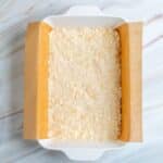 A square white baking dish lined with parchment paper and filled with an even layer of unbaked crumbly dough mixture on a marbled surface.