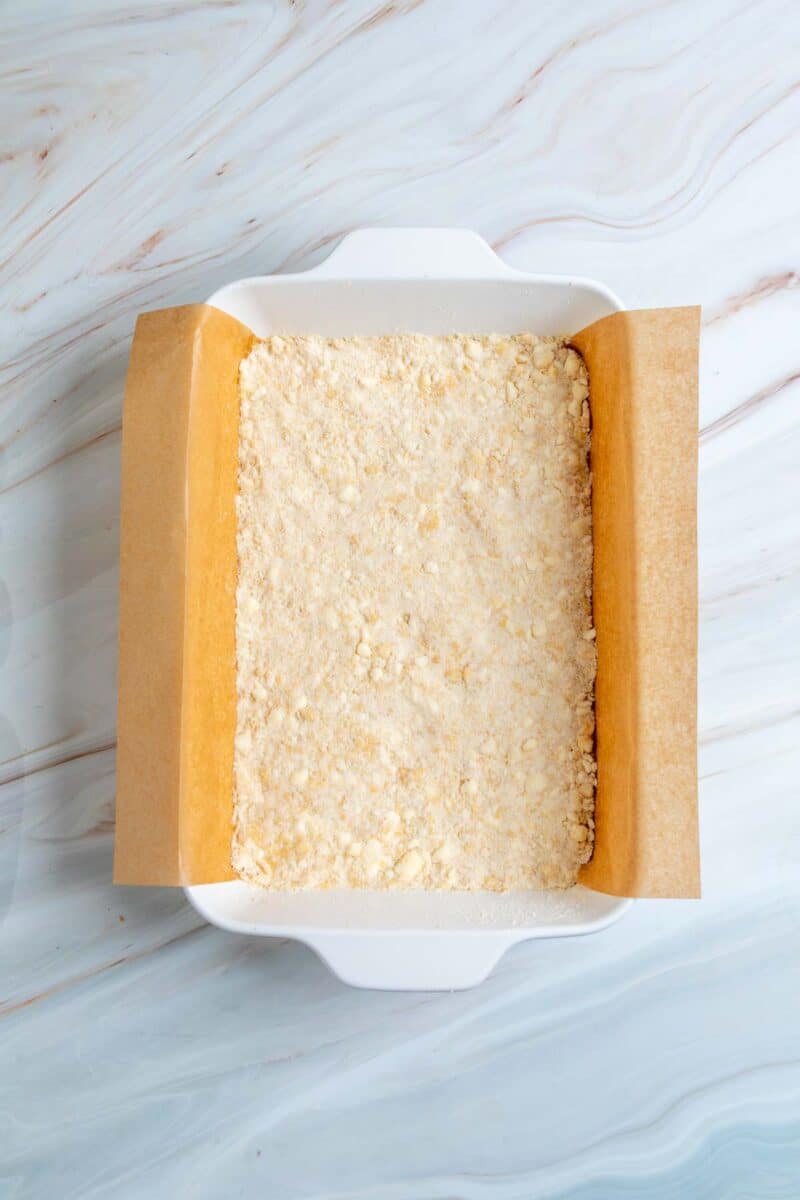 A square white baking dish lined with parchment paper and filled with an even layer of unbaked crumbly dough mixture on a marbled surface.