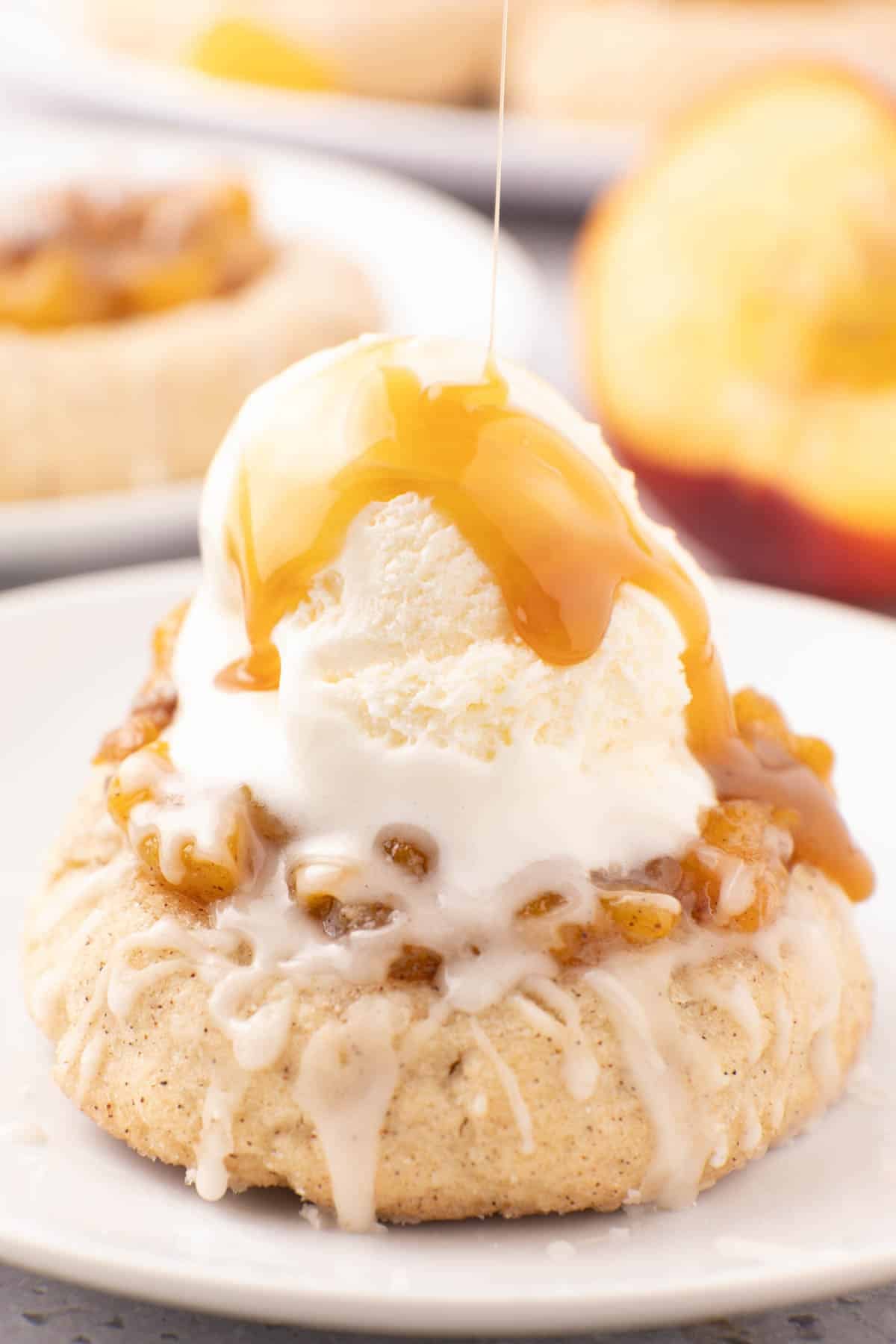 A peach cobbler cookie topped with vanilla ice cream and fruit, being drizzled with caramel sauce, is served on a white plate.
