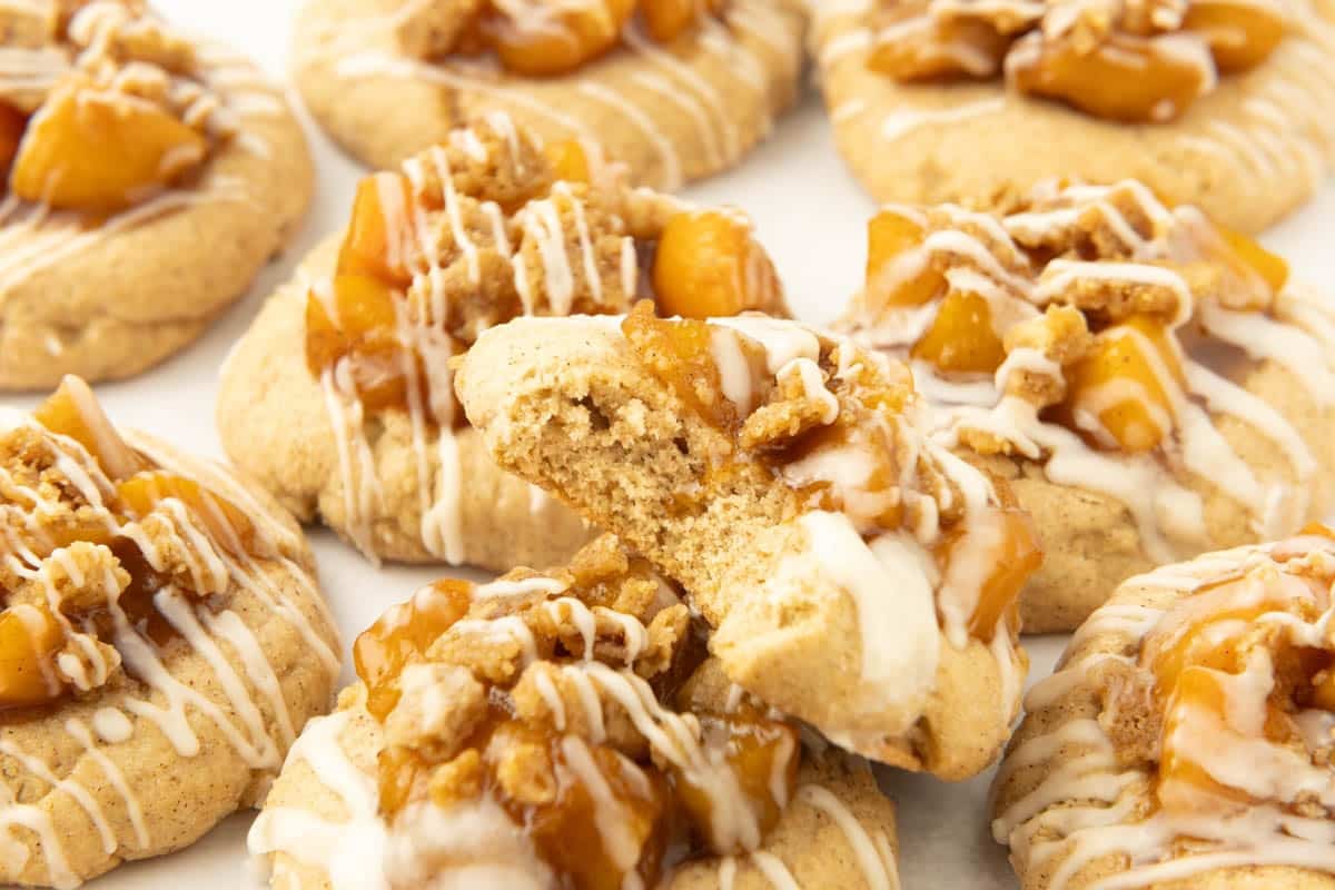 A group of peach cobbler cookies topped with apple chunks, crumble, and white icing, with one cookie in the center broken in half to show the inside.
