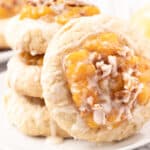 Three stacked peach cobbler cookies topped with peach filling and white icing sit on a white plate, with another cookie and a peach blurred in the background.
