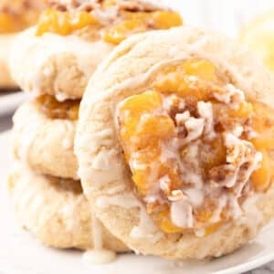 Three stacked peach cobbler cookies topped with peach filling and white icing sit on a white plate, with another cookie and a peach blurred in the background.