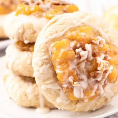 Three stacked peach cobbler cookies topped with peach filling and white icing sit on a white plate, with another cookie and a peach blurred in the background.