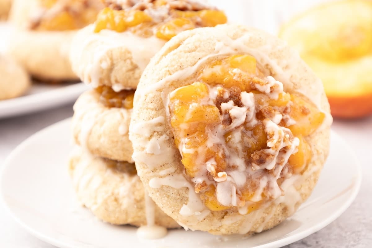 Three stacked peach cobbler cookies topped with peach filling and white icing sit on a white plate, with another cookie and a peach blurred in the background.