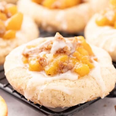 A close-up of a baked peach cobbler cookie topped with diced apples, cinnamon crumble, and white icing, resting on a black cooling rack.