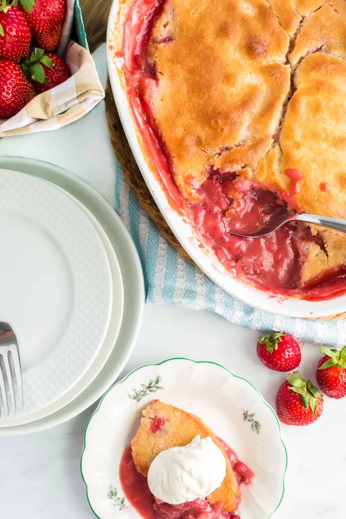 Baked strawberry cobbler in a dish with a serving on a plate, topped with vanilla ice cream. Fresh strawberries and empty plates are nearby.