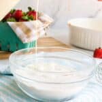 A bowl of dry baking ingredients is being mixed with liquid as someone pours vanilla extract from a measuring spoon; strawberries and kitchen items are in the background.