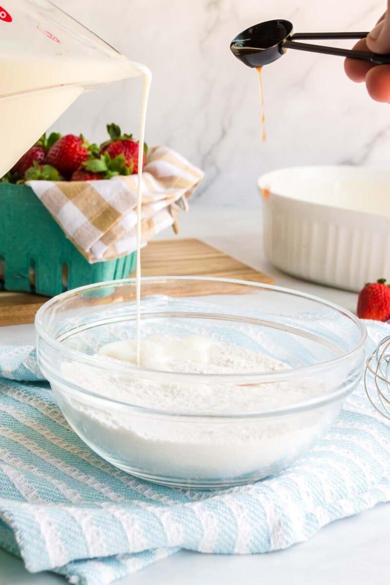 A bowl of dry baking ingredients is being mixed with liquid as someone pours vanilla extract from a measuring spoon; strawberries and kitchen items are in the background.