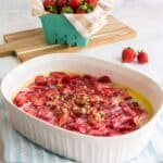 White baking dish with sliced strawberries and crumb topping on a striped towel, with fresh strawberries in a basket and cutting board in the background.