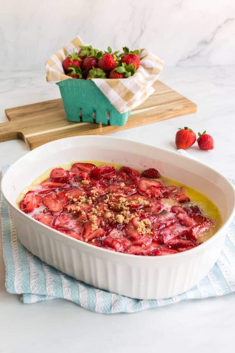 White baking dish with sliced strawberries and crumb topping on a striped towel, with fresh strawberries in a basket and cutting board in the background.