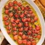 Oval dish filled with tomato confit, garnished with herbs. Slices of bread are placed around the dish on a wooden surface.